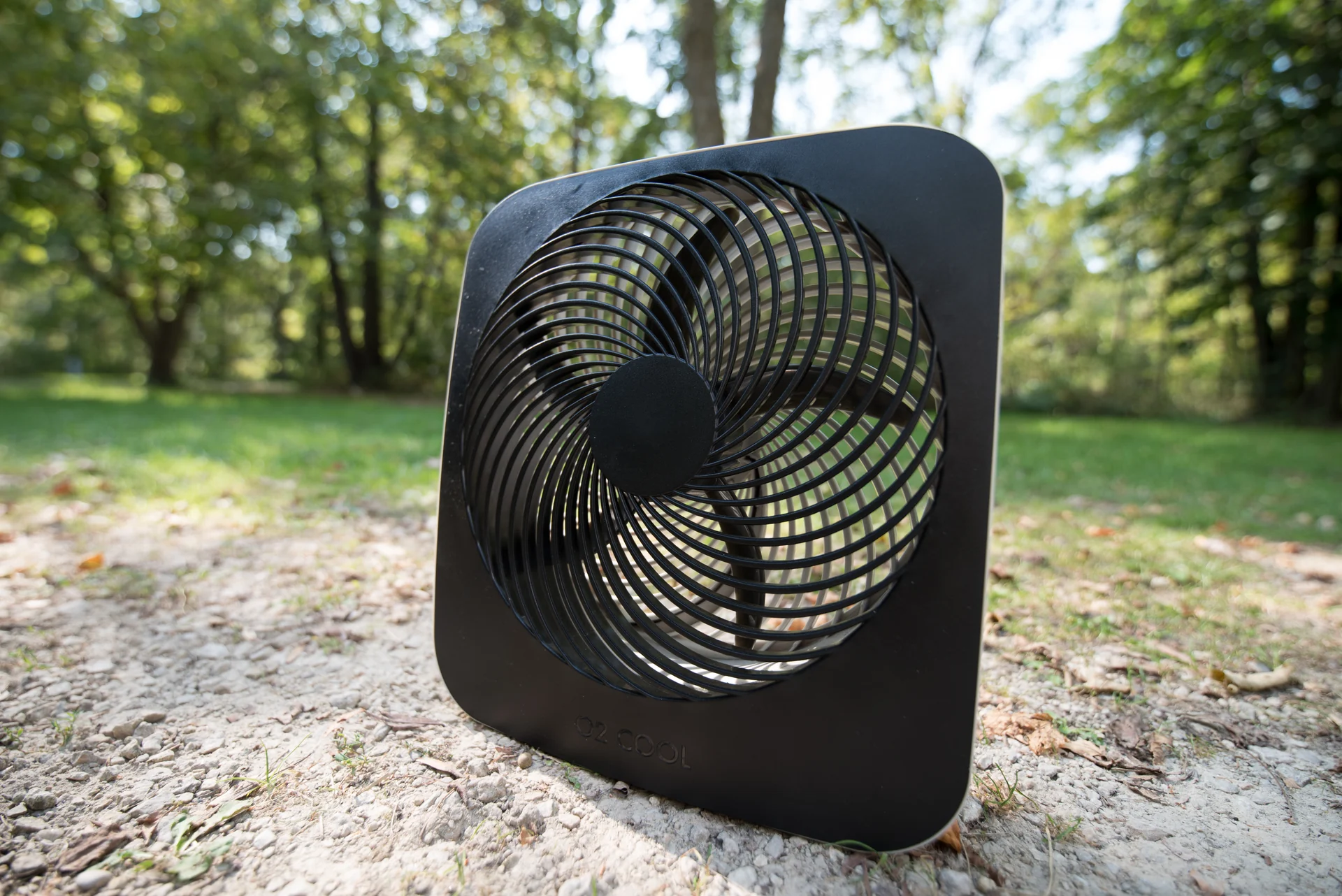 露營營地地面上的黑色 O2COOL 10 吋電池式風扇，背景為林地與草地